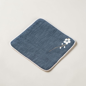 Puede incluir: Un pa&ntilde;o de lavado cuadrado, azul marino, con un borde blanco. Un delicado dise&ntilde;o floral con flores blancas y tallos dorados est&aacute; bordado en la esquina. El pa&ntilde;o est&aacute; hecho de una tela texturizada.
