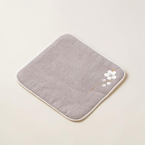 Puede incluir: Un pa&ntilde;o cuadrado gris con un borde blanco y un dise&ntilde;o floral en la esquina. El dise&ntilde;o floral presenta una flor blanca con detalles dorados. El pa&ntilde;o est&aacute; hecho de una tela texturizada.