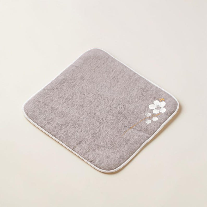 Puede incluir: Un pa&ntilde;o cuadrado gris con un borde blanco y un dise&ntilde;o floral en la esquina. El dise&ntilde;o floral presenta una flor blanca con detalles dorados. El pa&ntilde;o est&aacute; hecho de una tela texturizada.