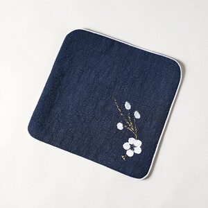 Puede incluir: Pa&ntilde;o cuadrado azul marino con borde blanco. Un dise&ntilde;o floral con p&eacute;talos blancos y detalles dorados est&aacute; pintado sobre el pa&ntilde;o. El pa&ntilde;o es probablemente un art&iacute;culo decorativo o una toalla peque&ntilde;a.