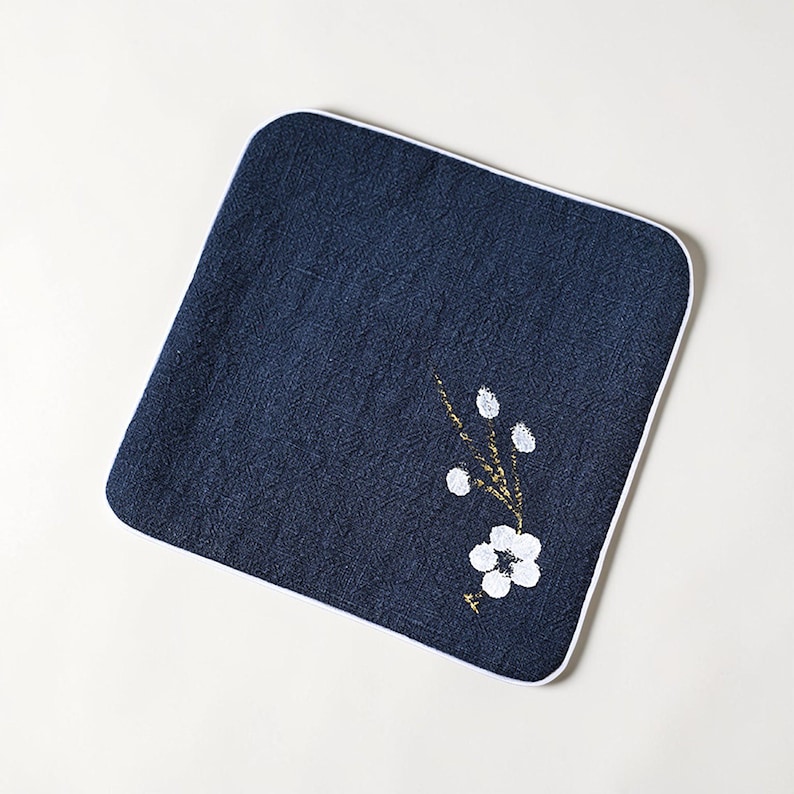 Puede incluir: Pa&ntilde;o cuadrado azul marino con borde blanco. Un dise&ntilde;o floral con p&eacute;talos blancos y detalles dorados est&aacute; pintado sobre el pa&ntilde;o. El pa&ntilde;o es probablemente un art&iacute;culo decorativo o una toalla peque&ntilde;a.