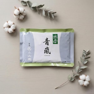 Könnte beinhalten: Eine Packung Matcha-Tee mit japanischen Schriftzeichen und dem Wort "AOARASHI" in Schwarz. Die Packung ist weiß mit grauen und grünen Akzenten. Baumwollknospen und Eukalyptuszweige sind um die Packung herum auf einer hellen Holzoberfläche angeordnet.