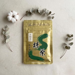 May include: A gold-colored package of matcha tea with Japanese text and a large green brushstroke design. The package also includes the text "100g" and "MARUKYU KOYAMAEN". The image is styled with sprigs of eucalyptus and a cotton flower.
