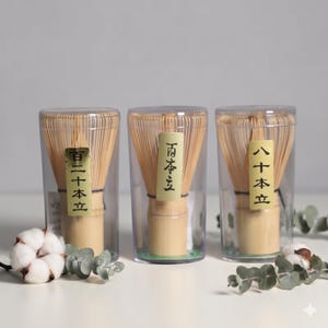 May include: Three bamboo matcha whisks in clear plastic containers. Each whisk has a light brown handle and numerous thin tines. The containers have labels with Japanese text. Eucalyptus and cotton accents are in the foreground.