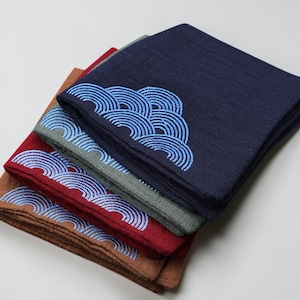 May include: A stack of four folded square handkerchiefs in shades of brown, burgundy, gray, and navy blue. Each handkerchief features a blue embroidered wave pattern in the corner. The fabric has a textured appearance.