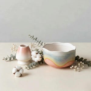 Puede incluir: Un cuenco de matcha de cerámica con borde ondulado, con un degradado de colores pastel: rosa, amarillo y azul. También hay un soporte para batidor de matcha de cerámica a juego con un degradado rosa y blanco. La escena está decorada con algodón y vegetación.