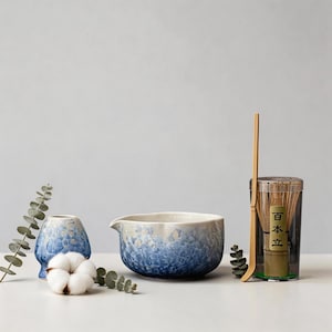 Puede incluir: Naturaleza muerta con un cuenco de té de cerámica azul y blanco y un jarrón pequeño. También hay un batidor de bambú y un recipiente de té matcha. Los artículos están dispuestos con ramitas de vegetación y una bola de algodón.