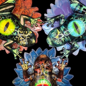 May include: Three colorful frog sculptures with large, detailed eyes and intricate patterns. Each frog is positioned on a stylized flower, with vibrant hues of green, orange, blue, and gold. The background is black, highlighting the sculptures.