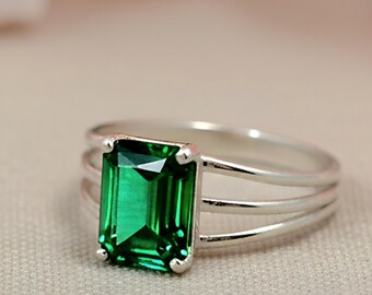 May include: A silver ring featuring a large, emerald-cut, vibrant green gemstone. The ring has a triple-band design, with the gemstone set prominently in the center. The ring is displayed against a soft, neutral background, with blurred pink flowers in the background.