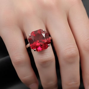 Może przedstawiać: Srebrny pierścionek z dużą, kwadratową, czerwoną szlachetnym kamieniem. Pierścionek jest pokazany na dłoni, a kamień szlachetny odbija światło. Tło jest ciemne.