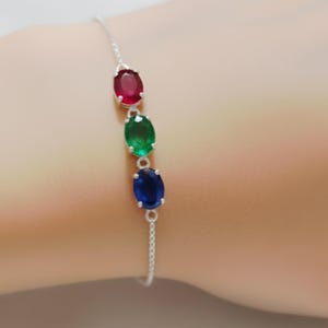 May include: A silver bracelet with three oval gemstones in red, green, and blue. The gemstones are set in a row along the delicate chain. The bracelet is displayed on a person's wrist.