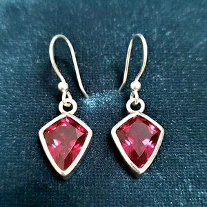 May include: A pair of silver drop earrings with a geometric design. Each earring features a faceted, deep red gemstone set in a silver bezel. The gemstones are diamond-shaped. The earrings hang from a simple silver hook.