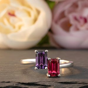 May include: Two silver rings, each featuring a rectangular gemstone. One ring has a deep purple stone, the other a vibrant red stone. The rings are set against a dark background with soft, blurred flowers in the background.