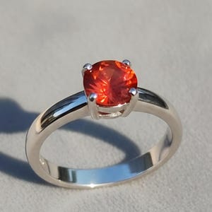 Peut inclure: Une bague en argent ornée d'une pierre précieuse orange vif et ronde. La pierre est sertie de quatre griffes, et le corps de la bague est lisse et poli. La bague est présentée sur une surface gris clair, avec une ombre portée à gauche.