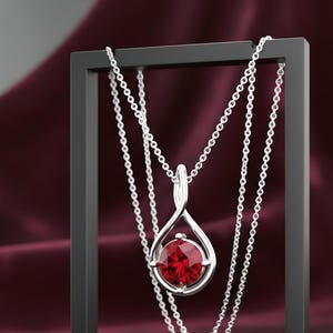 May include: A silver pendant necklace with a large, round, red gemstone. The pendant hangs from a delicate chain and is displayed on a black stand against a burgundy background. The necklace is a piece of jewelry.