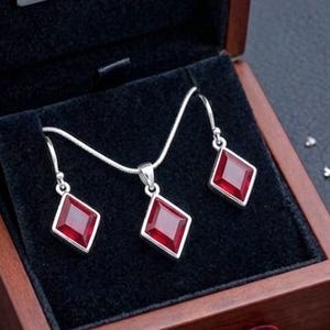 May include: A jewellery set in a wooden box. The set includes a necklace and earrings with red, diamond-shaped gemstones. The box is open, revealing the jewellery on a black velvet lining. The box has the text "GEMME ÉLÉGANTE".
