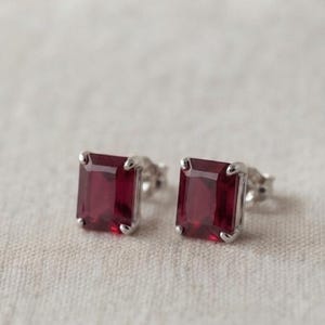 May include: A pair of stud earrings featuring rectangular, emerald-cut deep red gemstones set in polished silver-toned metal prongs. Each earring has a clear, faceted surface that reflects light, showcasing the vibrant crimson colour of the stones.