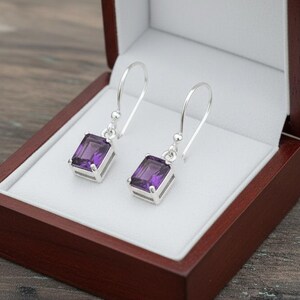 May include: Silver drop earrings featuring rectangular, deep purple gemstones. The earrings are presented in an open, dark wood jewellery box with a white interior. The box rests on a wooden surface.