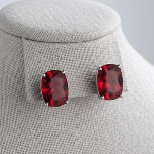 May include: A pair of stud earrings featuring deep red, cushion-cut gemstones set in silver-toned metal prongs. The stones have a faceted surface that reflects light, and each earring is secured with a post back mechanism for attachment.