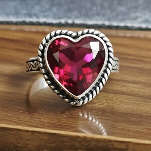 May include: A sterling silver ring with a large, faceted, heart-shaped, ruby-red gemstone. The ring's setting features a twisted rope design around the heart and decorative detailing on the band.