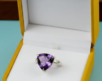May include: A silver ring with a large, triangular, deep purple gemstone is displayed in an open yellow velvet jewelry box with a white interior. The ring is centered in the box, showcasing the gemstone's facets.
