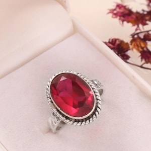 May include: A silver ring featuring a large, oval, ruby-red gemstone. The ring has a rope-style border around the stone and is presented in a white jewellery box. The ring is a classic design.