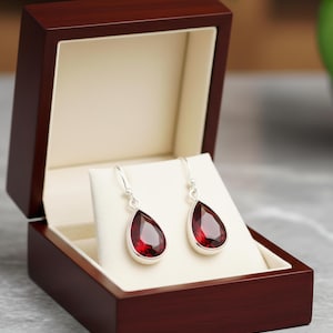 May include: A pair of teardrop-shaped earrings with a deep red gemstone set in a silver metal frame. The earrings are displayed in an open, dark wood jewelry box with a cream-colored interior. The earrings are on a white display.