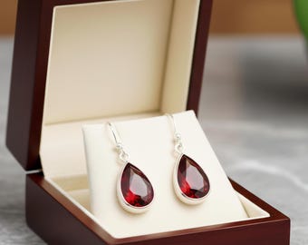 May include: Teardrop earrings with a rich red gemstone, set in silver. The earrings are presented in an open, dark wood jewellery box with a cream interior. The earrings are displayed on a white stand.