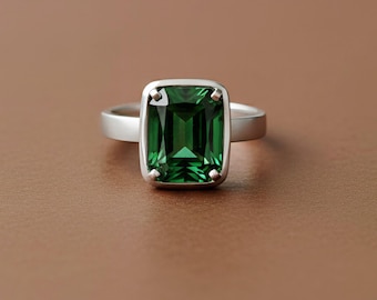May include: A silver ring featuring a large, emerald-cut, deep green gemstone. The gemstone is set in a square bezel setting, and the band is a simple, polished silver. The ring is displayed on a brown surface.