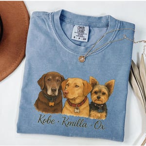 May include: A blue short-sleeved t-shirt featuring a graphic illustration of three dogs: a brown Labrador, a yellow Labrador, and a Yorkshire Terrier. Below the dogs, the names Kobe, Kmilla, and Ox are printed in a dark cursive font.