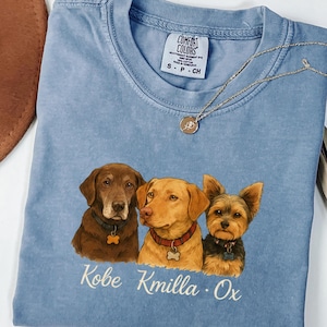 May include: A blue short-sleeved t-shirt featuring a graphic of three dogs wearing collars with bone-shaped tags. Below the dogs, the names Kobe, Kmilla, and Ox are printed in white script. The shirt has a Comfort Colors label at the neckline.