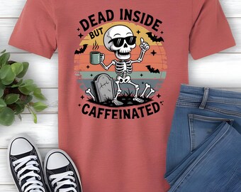 May include: A coral-colored t-shirt featuring a skeleton wearing sunglasses, holding a coffee mug, and giving a thumbs-up. The shirt has the text "DEAD INSIDE BUT CAFFEINATED" with bats and a sunset graphic. Black and white sneakers and blue jeans are also visible.