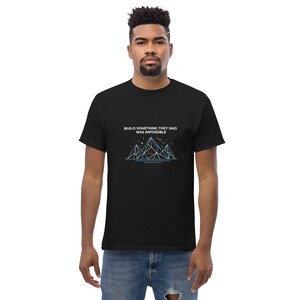 Op de afbeelding: Zwart T-shirt met de tekst "BUILD SOMETHING THEY SAID WAS IMPOSSIBLE" en een lichtblauw geometrisch bergontwerp. Het T-shirt heeft een ronde hals.