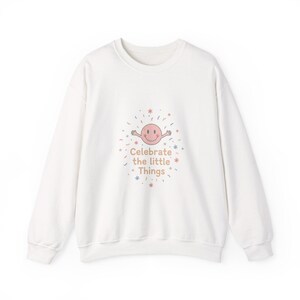 Könnte beinhalten: Weißes Sweatshirt mit Rundhalsausschnitt. Auf der Vorderseite befindet sich eine Grafik mit einem lächelnden Gesicht und dem Text "Celebrate the little Things". Das Design umfasst bunte Konfetti und Sternakzente.