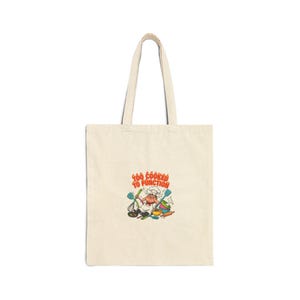 Puede incluir: Bolso tote de color crema con un gráfico de chef de dibujos animados y el texto "Too Cooked to Function" en naranja y amarillo. El chef lleva un gorro blanco y sostiene utensilios de cocina.