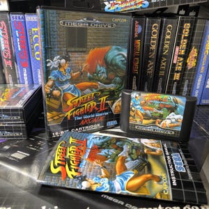 Pode incluir: Uma coleção de cartuchos e caixas de videojogos Sega Mega Drive, incluindo "Street Fighter II: The World Warrior" e "Golden Axe". A arte da caixa de "Street Fighter II" mostra uma mulher com um vestido azul lutando contra um personagem de pele verde. O cartucho do jogo também é visível.