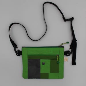 May include: Green crossbody bag with a patchwork design. The bag features a black adjustable strap, a zippered main compartment, and a smaller zippered pocket. The bag has a black and grey colour block design with a small button closure.