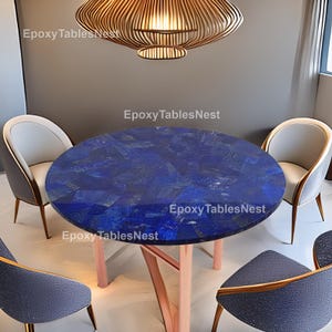 Puede incluir: Una mesa de comedor redonda con una superficie azul oscuro, estilo mosaico. La mesa está sostenida por patas de color rosa claro. La imagen también muestra cuatro sillas con asientos grises y respaldos de color claro. El texto "EpoxyTablesNest" es visible.