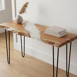 Può includere: Una consolle con bordo vivo, finitura in legno chiaro e un fiume di resina bianca. Il tavolo ha gambe a forcina nere ed è sormontato da una pila di libri e un piccolo vaso con erba secca.
