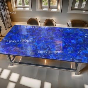 May include: A rectangular table with a vibrant blue mosaic top. The table's surface is composed of irregularly shaped pieces, creating a textured appearance. The base is made of silver metal. The text "Epoxy Tables Nest" is visible on the table.