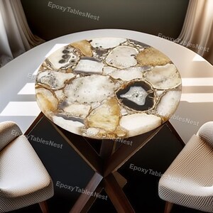 May include: A circular table with a natural stone top, showcasing a mosaic of beige, white, and black sections. The table is supported by a dark wooden base. The text "Epoxy Tables Nest" is visible.