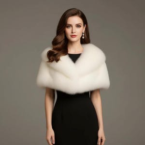 Puede incluir: Un chal de piel sint&eacute;tica blanco con un cuello ancho, sobre un vestido negro. El chal tiene una textura suave y esponjosa. La mujer de la imagen lleva pendientes colgantes.
