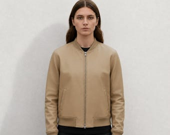 Puede incluir: Una chaqueta tipo bomber de color beige con cremallera metálica plateada de longitud completa, cuello, puños y dobladillo acanalados. La chaqueta cuenta con dos bolsillos verticales ribeteados en la parte delantera y se lleva sobre una camiseta negra.