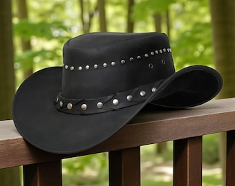 Puede incluir: Un sombrero de vaquero negro con ala ancha y copa redondeada. El sombrero está adornado con tachuelas plateadas a lo largo de la corona y una banda de cuero trenzado con tachuelas plateadas. El sombrero descansa sobre una barandilla de madera marrón.
