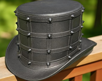May include: A black leather top hat with a studded design. The hat features a cylindrical crown with a grid pattern and is adorned with silver-colored spikes. The brim is flat and the hat is sitting on a wooden surface.