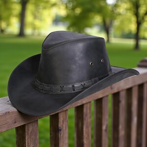 May include: A black leather cowboy hat with a wide brim and a decorative band around the crown. The hat has two small eyelets on the side and is resting on a wooden railing. The background is a blurred view of green trees.