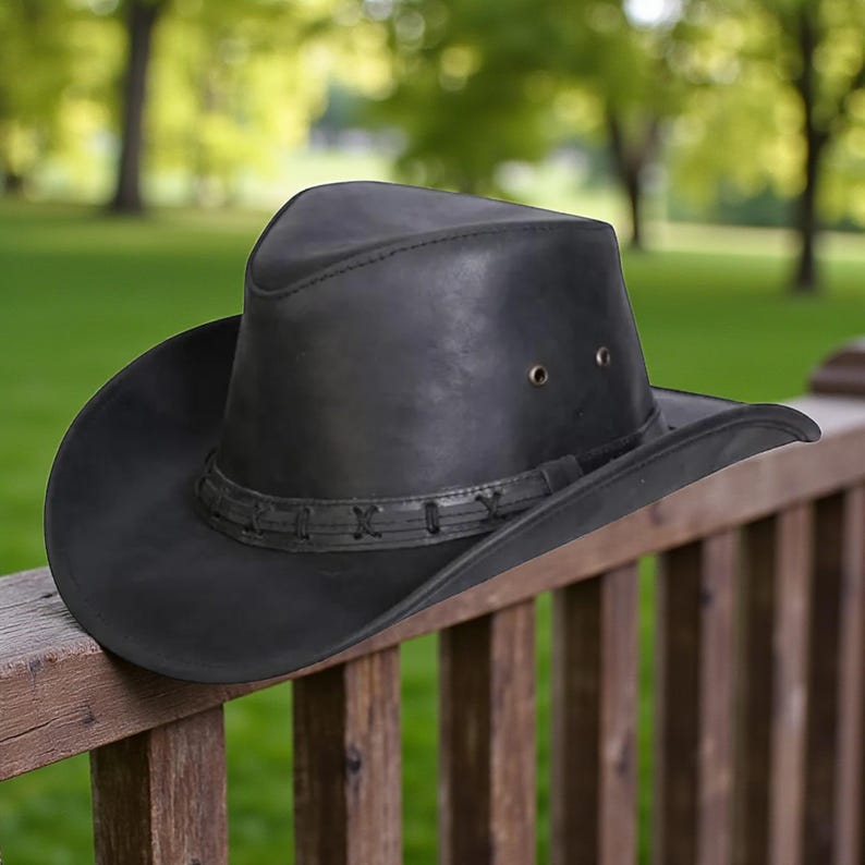 May include: A black leather cowboy hat with a wide brim and a decorative band around the crown. The hat has two small eyelets on the side and is resting on a wooden railing. The background is a blurred view of green trees.