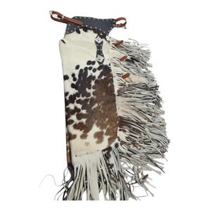 May include: Cowhide chaps with a brown and white spotted pattern. The chaps have a black leather top with silver-coloured embellishments and long white fringe along the sides. Brown leather straps and buckles are visible.