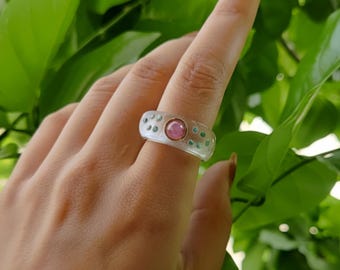 May include: A clear ring with a pink gemstone center and small green and white accents. The ring is on a finger, set against a backdrop of green leaves.