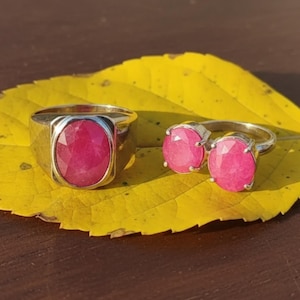 May include: Two rings with large, oval, pink gemstones. One ring has a single stone set in a square band. The other ring has two stones set side-by-side. The rings rest on a yellow leaf.